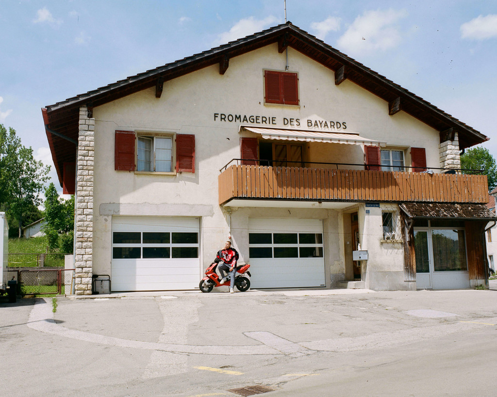 An image from the collection “CH 2127 Les Bayards” by Bryan Colò, who takes a satirical angle to talk about his origins. Here, the model is shot in front of the local dairy - © Photograph: Cynthia Mai Amman, Swiss Design Awards Blog